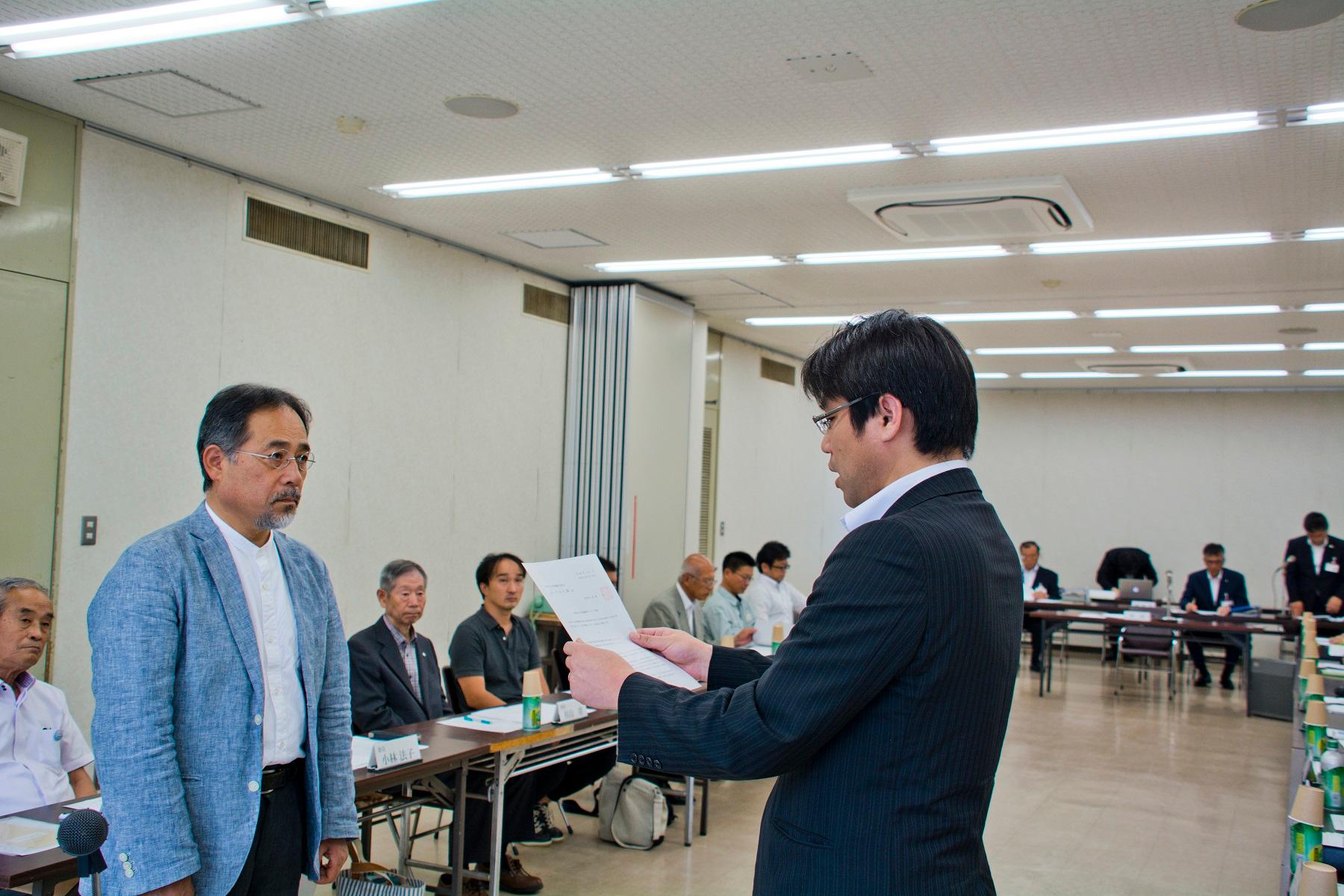 会議室内で多くの人が見守る中、右側の黒いスーツの男性が左側の青いジャケットの男性に書類をわたす写真