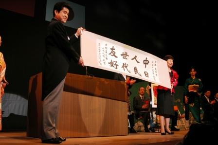 合併1周年記念式典で「中日人民世代友好」と書かれた記念品を持っている陳愛莉棗荘市副市長と燕尾服を着た市長の写真