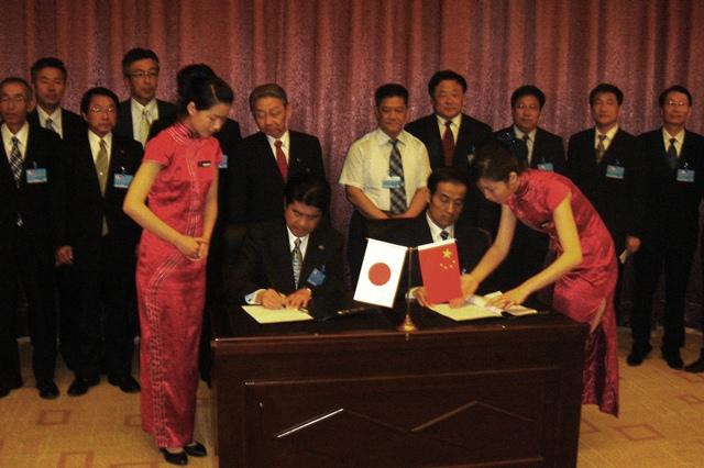 村田弘司 美祢市長と張宝民 棗荘市副市長が机に座り、合意書に署名しており、その様子を見ている関係者の方々の写真