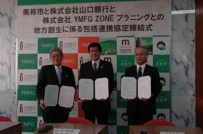 美祢市市長と株式会社山口銀行、株式会社YMFG ZONEプラニングの関係者の方々が調印した文書を手に持っている写真