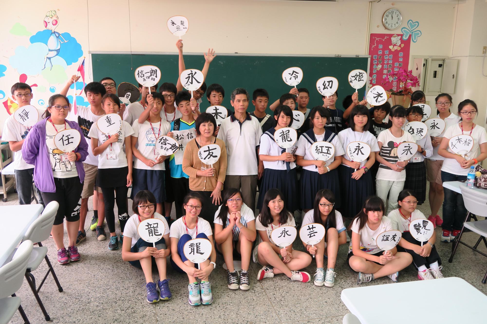 大人数の男女の中学生が教室で記念撮影をしている写真。1人1枚うちわをもち、うちわにはそれぞれ異なる漢字が書かれている。